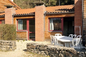 Una cabaña de madera con techo de tejas, pared de piedra y una mesa blanca con sillas afuera.