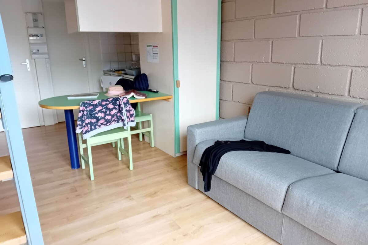 Una habitación con un sofá gris, una mesa verde con un libro y un sombrero, y piso de madera.