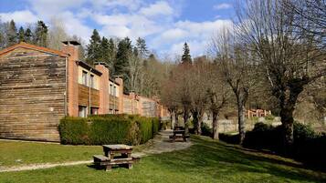 A row of wooden buildings with a paved path and benches, surrounded by trees and greenery.