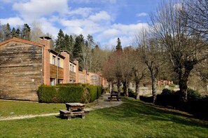 Une rangée de bâtiments en bois, avec un chemin pavé et des bancs, le tout entouré d’arbres et de verdure.