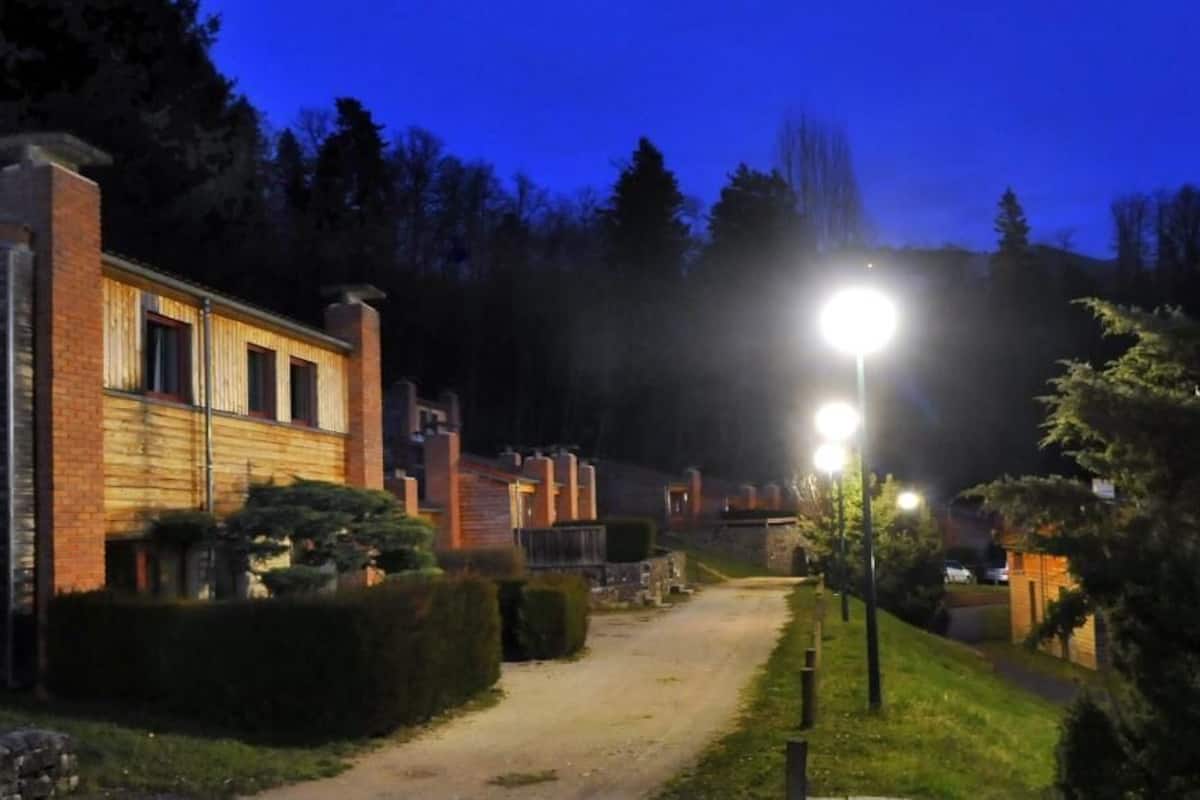 A pathway leading to a building with a brick facade and wooden siding, surrounded by greenery and illuminated by street lamps.