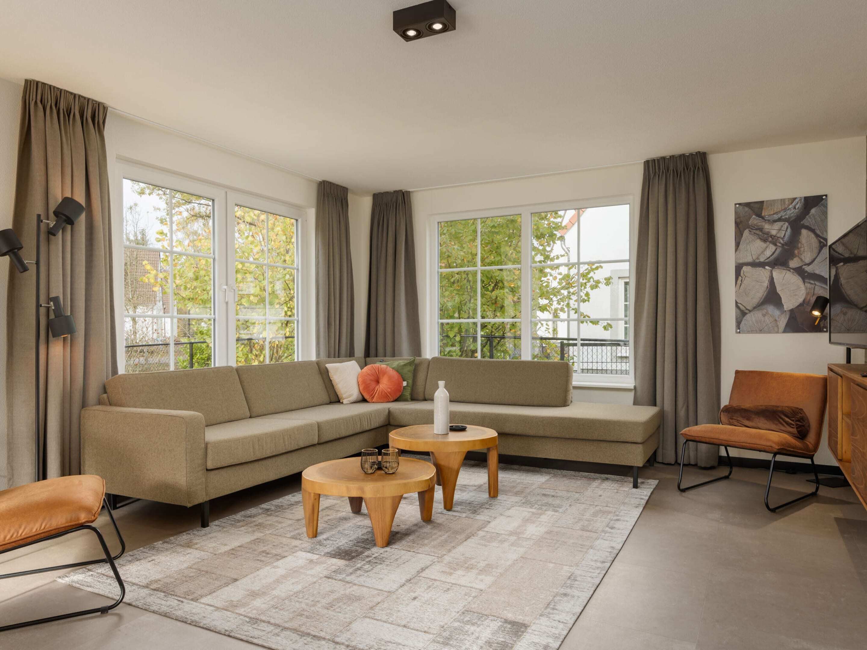 Ein modernes Wohnzimmer mit einem Ecksofa, zwei Holztischen, einem Ledersessel und großen Fenstern mit Vorhängen.