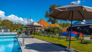 Ein Schwimmbecken mit Liegestühlen und Sonnenschirmen, umgeben von einer Rasenfläche und Gebäuden mit orangefarbenen Dächern.