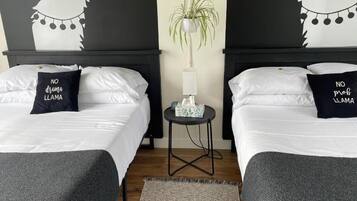 A hotel room with two beds, each with a pillow that reads "NO drama llama." A small table with a plant and a lamp is between the beds.