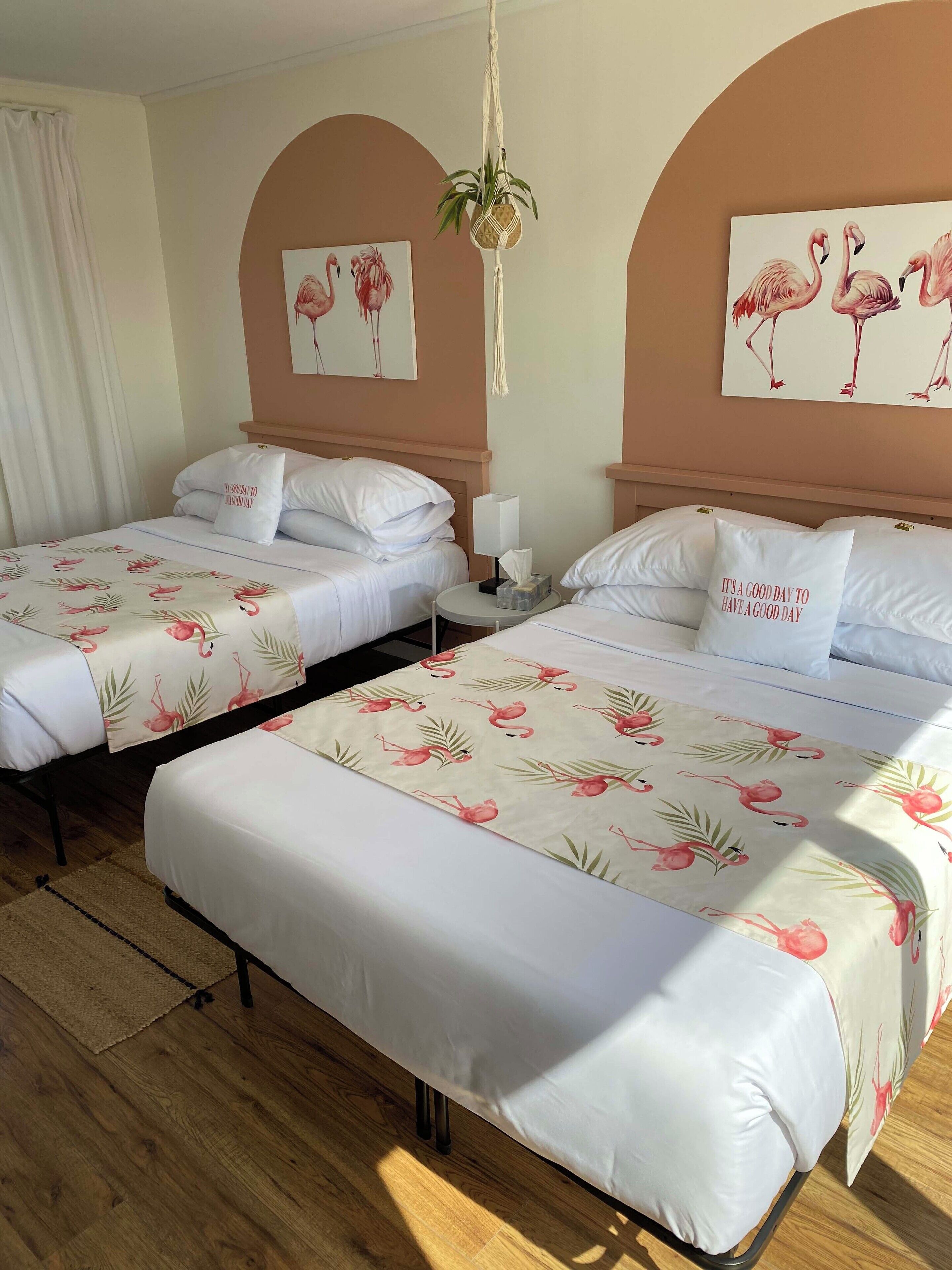 Habitació d'hotel amb dos llits, cadascun amb llençols blancs, coixins a joc i una col·cha amb estampat floral. Hi ha una obra d'art a la paret amb flamencs, una planta penjant i una taula de nit amb una làmpada i una caixa de mocadors.