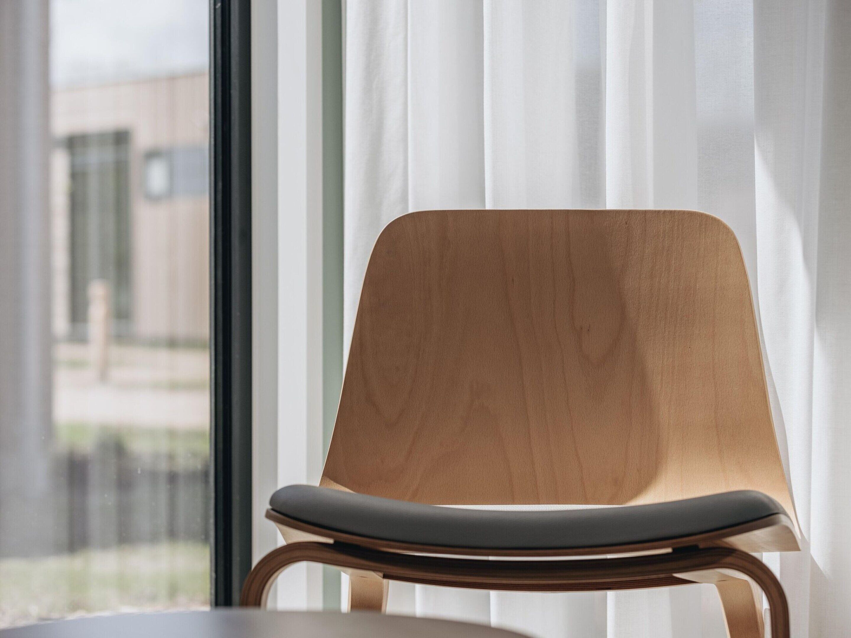 A modern chair with a wooden backrest and a cushioned seat, positioned in front of a window with sheer curtains.