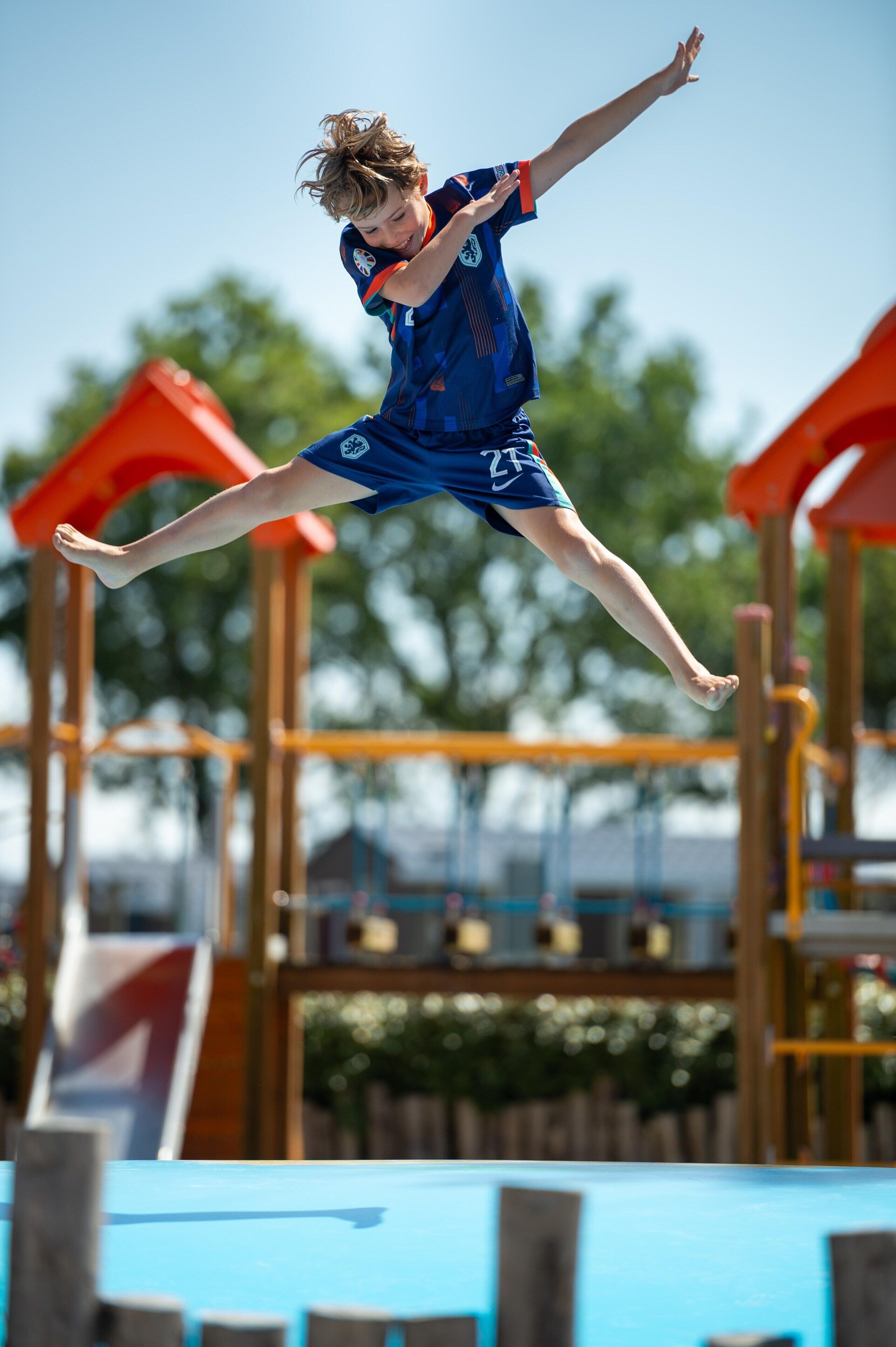 Un enfant saute sur un trampoline dans un espace de jeux extérieur, équipé de structures en bois et d’un toboggan.
