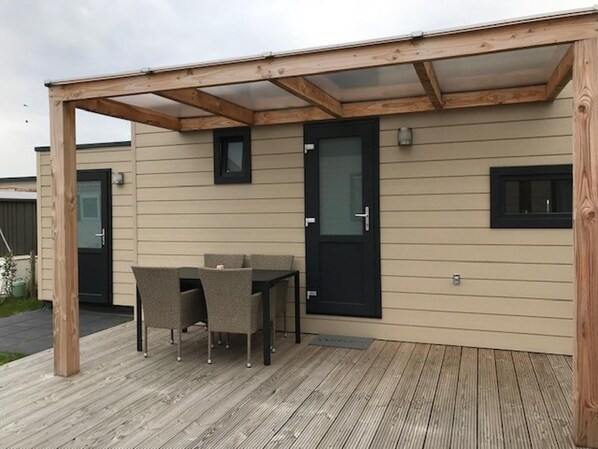 A covered outdoor area with a wooden deck, a table with chairs, and a door with a window. - Chalet in Netherlands With Veranda & Comfort (Koudekerke)