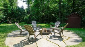 Un espacio circular con asientos, sillas Adirondack alrededor de un brasero, situado en un jardín con césped, una caseta de madera y un parque infantil al fondo.