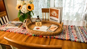 Una mesa de madera con un posavasos tejido, un jarrón con flores, una botella, un cartel de bienvenida y algunos folletos.