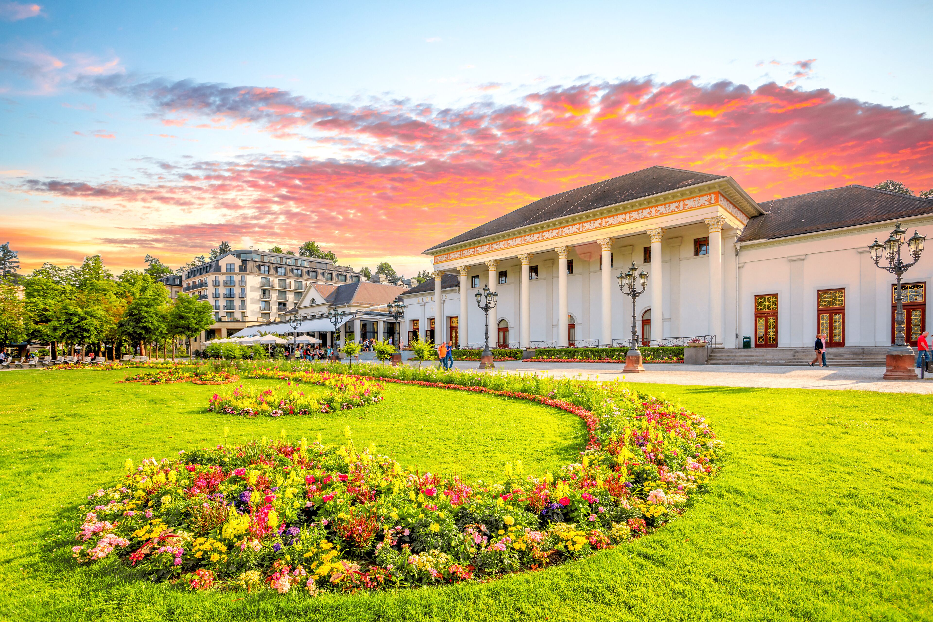 A historic building with classical architecture, surrounded by well-maintained gardens and a clear sky.