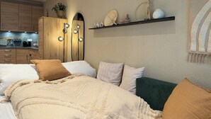Une chambre à coucher avec un lit, des oreillers, une table de chevet, une étagère avec des objets décoratifs, et un coin cuisine à l’arrière-plan.