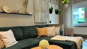Un canapé vert agrémenté de plusieurs coussins, une table basse en bois sur laquelle est posé un vase de fleurs, et une vue sur l’extérieur par une fenêtre.