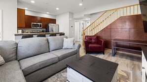 Ein modernes Wohnzimmer mit einem grauen Sofa, einem Couchtisch und einem roten Ledersessel. Im Hintergrund ist eine Küche mit Holzschränken und Edelstahlgeräten zu sehen.