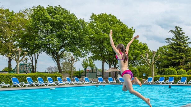 Una persona saltando a una piscina rodeada de sillas de descanso y árboles.