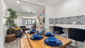 Un coin repas avec une table en bois dressée pour quatre personnes, entourée de chaises modernes. On peut apercevoir une grande plante en pot et un vase décoratif contenant des plumes d’herbe des pampas.