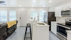 A modern kitchen with white cabinetry, a stainless steel refrigerator, a microwave, and an oven. There is a small island with a sink and a stool. - Hopkins (Biloxi)