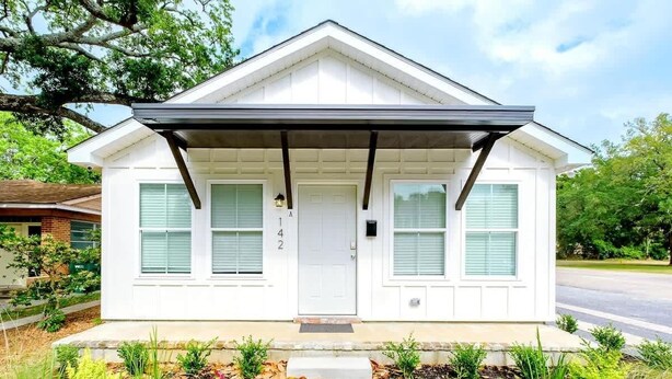 A white single-story house with a covered porch, a front door, and a small garden with various plants and flowers. - Hopkins (Biloxi)