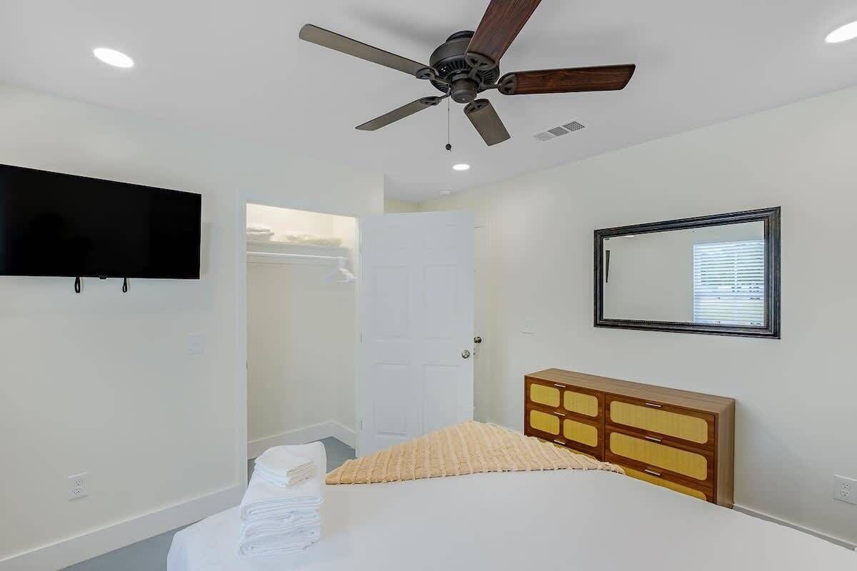 Un dormitorio con cama, ventilador de techo, televisión, espejo y ventana.
