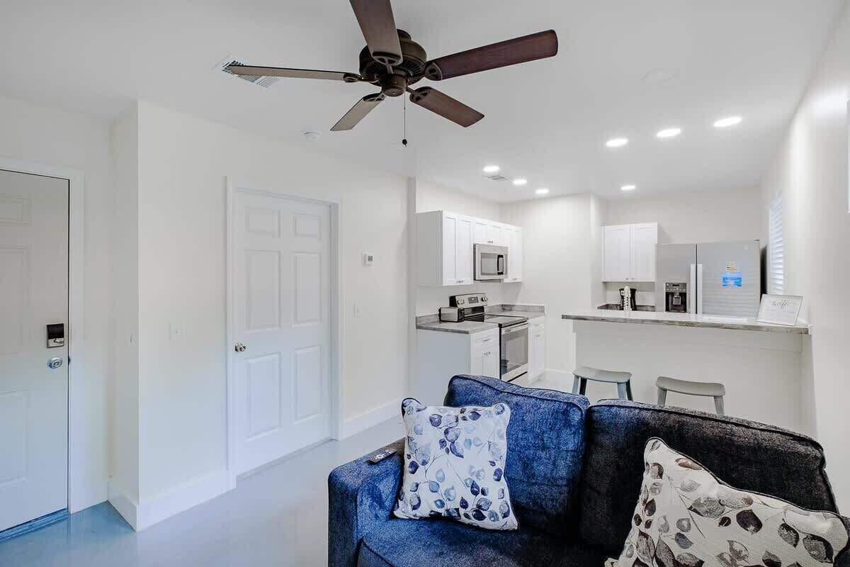 Un soggiorno moderno con ventilatore a soffitto, un divano con cuscini decorati e un angolo cottura con mobili e elettrodomestici bianchi.