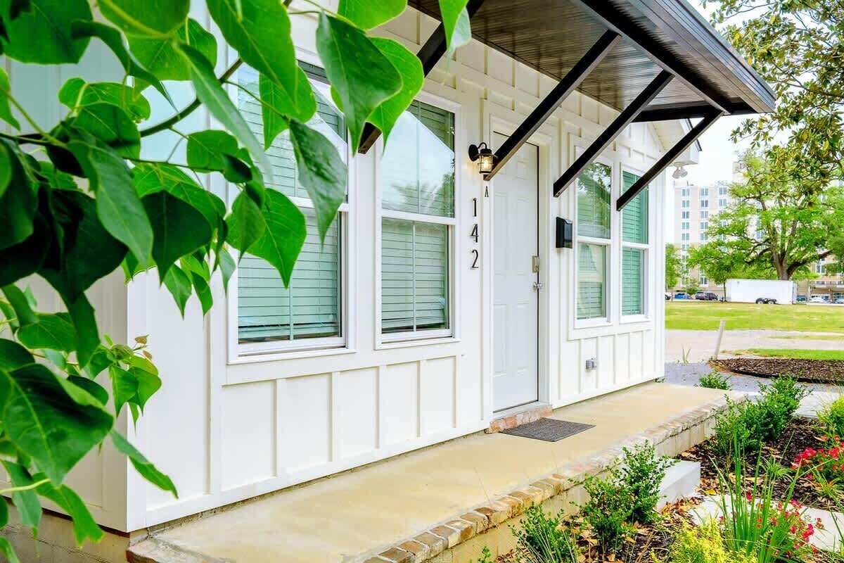 A white building with a number '142' on the door, a covered porch, and a garden with plants and flowers.