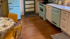 Een compacte keuken met een koelkast, fornuis, oven en vaatwasser. Er is een eettafel met stoelen en een raam met jaloezieën.