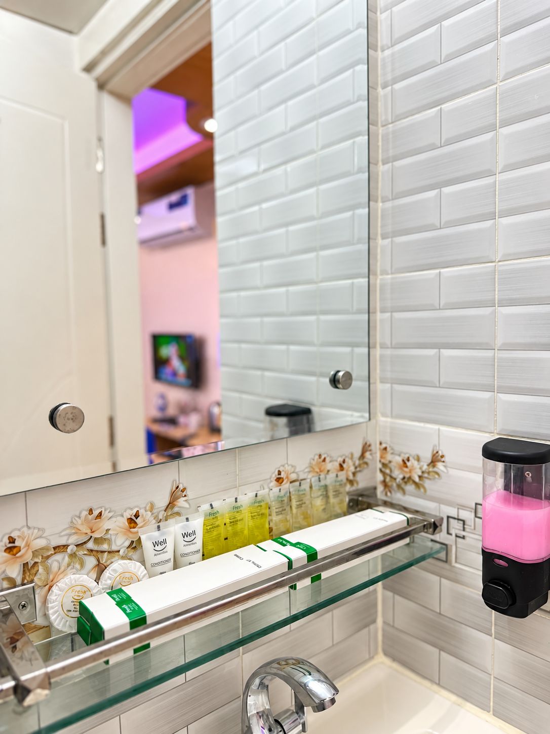 A bathroom with a glass shelf holding toiletries, a mirror, and a pink soap dispenser.