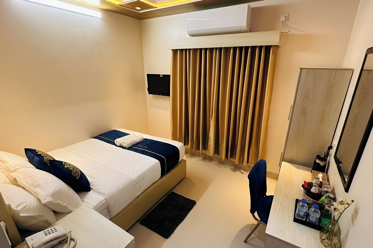 A hotel room with a bed, a desk, a chair, a mirror, a window with curtains, and an air conditioning unit.