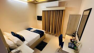 A hotel room with a bed, a desk, a chair, a mirror, a window with curtains, and an air conditioning unit.