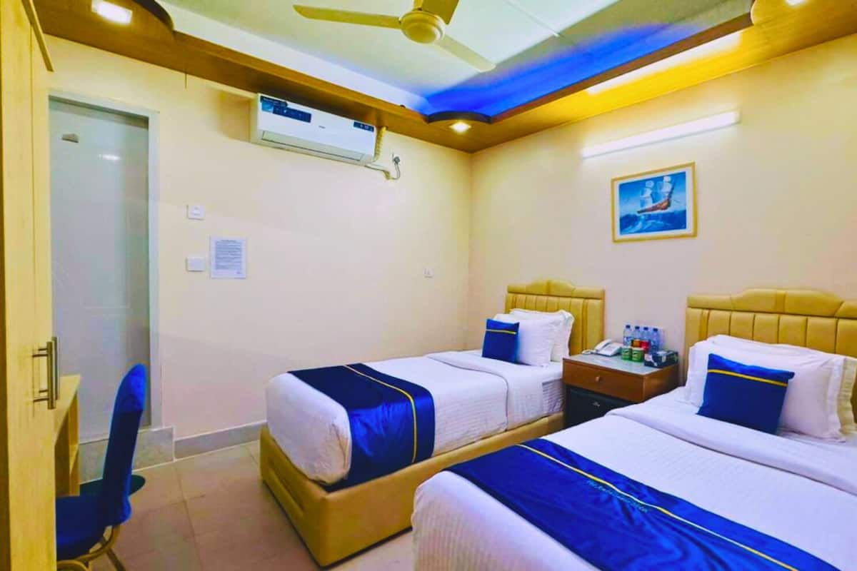 Una habitación de hotel con dos camas, un ventilador de techo, un equipo de aire acondicionado, una mesita de noche con un teléfono y botellas de agua, y un cuadro en la pared.