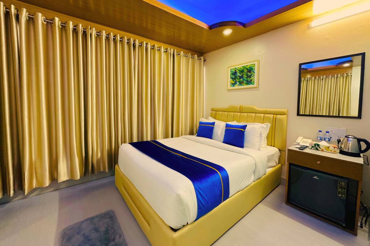 Habitación de hotel con cama, cortinas, mesita de noche con hervidor, espejo y un cuadro en la pared.