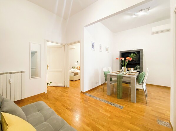 A modern living room with a wooden floor, a grey sofa, and a dining area with a table set for four. - Comfortable apartment located between Villa Borghese, Trinità dei Monti, and the Trevi Fountain (Roma)