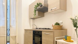 A modern kitchen with wooden cabinets, a built-in oven, a gas stove, and a small sink. There is a window with curtains, a white rug, and a small table with a vase and candles. - Comfortable apartment between Villa Borghese, Trinità dei Monti and Trevi Fountain (Roma)