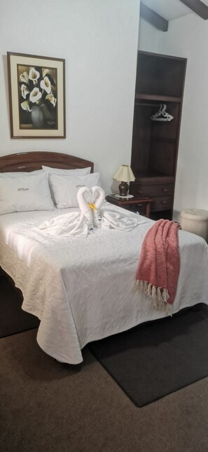 A neatly made bed with a heart-shaped towel arrangement, a framed floral artwork on the wall, a dark wooden wardrobe, and a small bedside table with a lamp. - EL CUMBE INN  (Cajamarca)