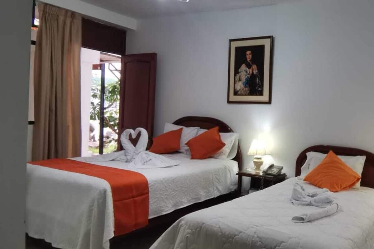 A hotel room with two beds, each with white bedding and orange pillows. There is a framed picture on the wall, a lamp on the bedside table, and a window with curtains.