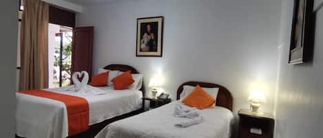 Habitación de hotel con dos camas, cada una con ropa de cama blanca y almohadas naranjas. En la pared, hay un cuadro enmarcado, una lámpara en la mesita de noche y una ventana con cortinas.
