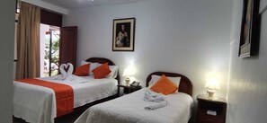 Habitación de hotel con dos camas, cada una con ropa de cama blanca y almohadas naranjas. En la pared, hay un cuadro enmarcado, una lámpara en la mesita de noche y una ventana con cortinas.