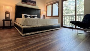 Une chambre spacieuse avec un grand lit, un plancher en bois, un plafond avec des poutres apparentes, une fenêtre donnant sur la verdure et un fauteuil près de la fenêtre.
