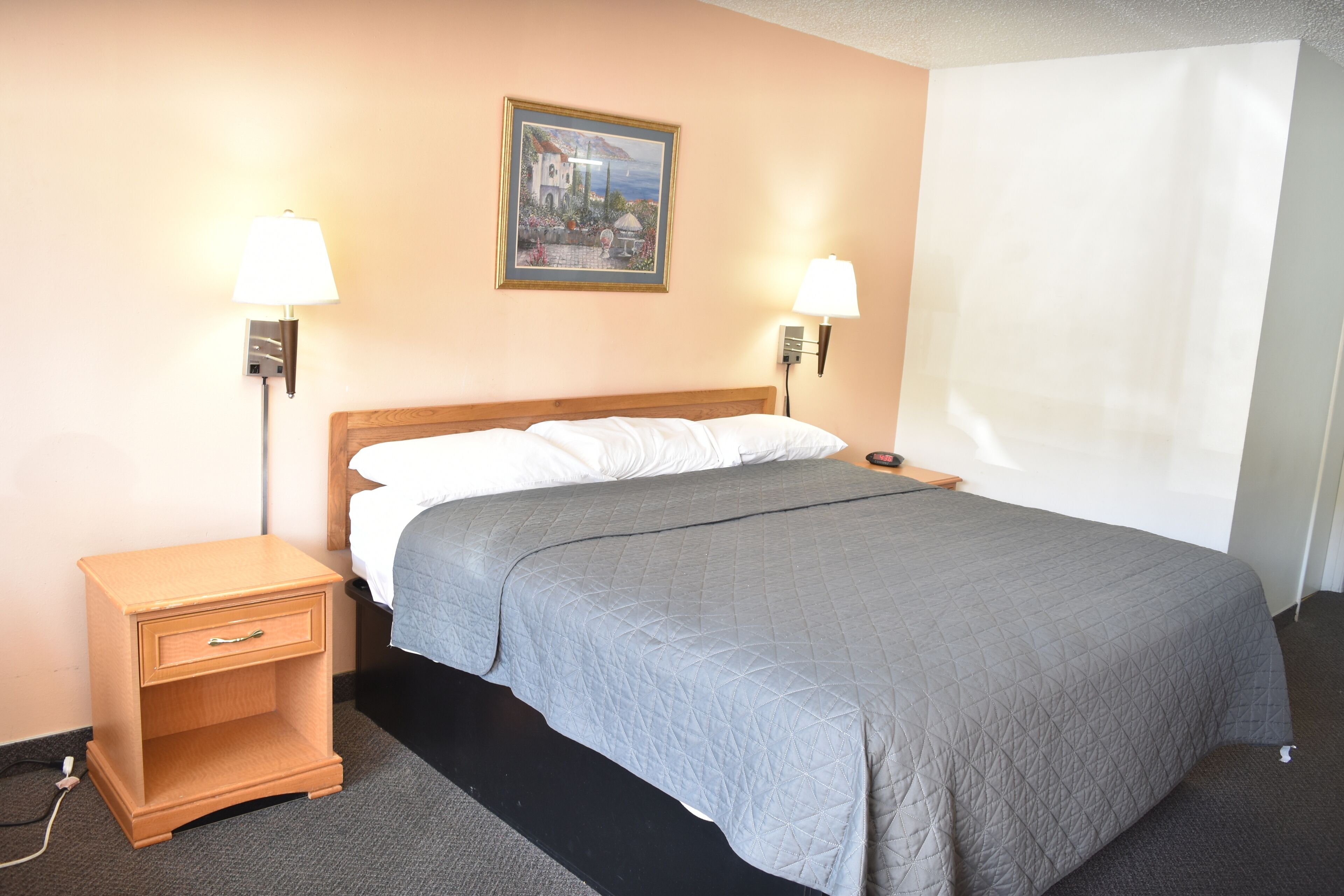 A hotel room with a bed, two bedside lamps, a nightstand with a drawer, a framed picture on the wall, and a window with a white curtain.