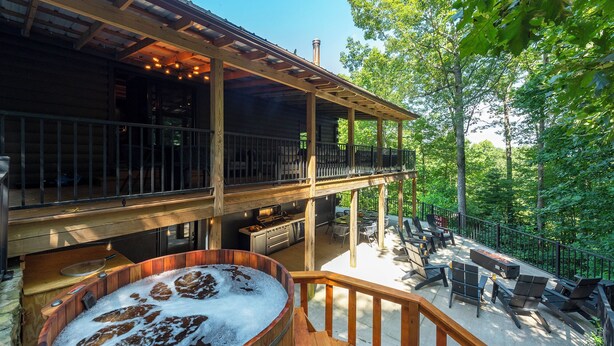 A wooden deck with a hot tub, outdoor kitchen, and seating area surrounded by trees.
