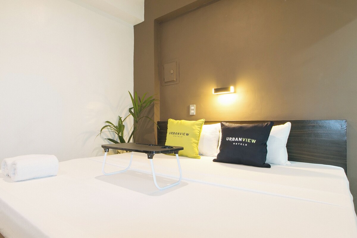 Habitación de hotel con cama, mesita de noche, lámpara y dos almohadas que llevan el nombre del hotel.