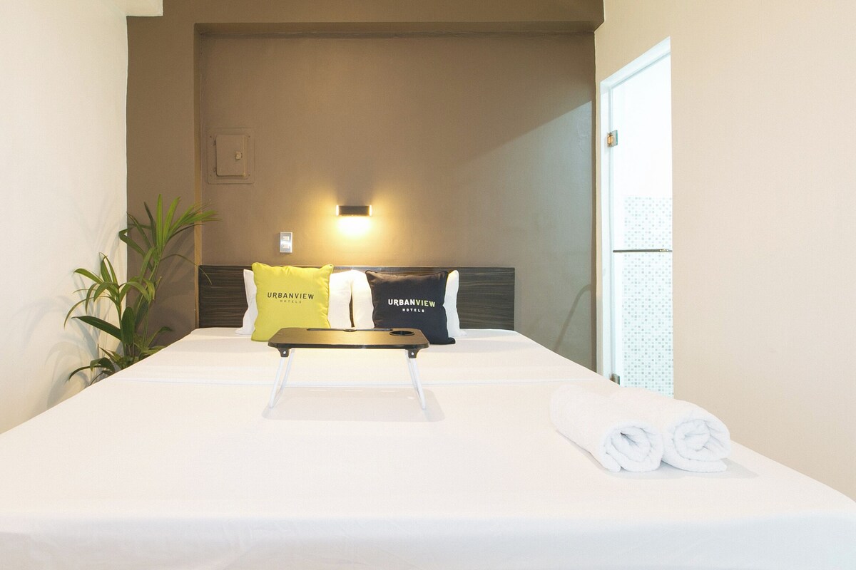 A hotel room with a bed, two pillows with "URBANVIEW HOTELS" branding, a bedside table, a small potted plant, and a wall-mounted light.