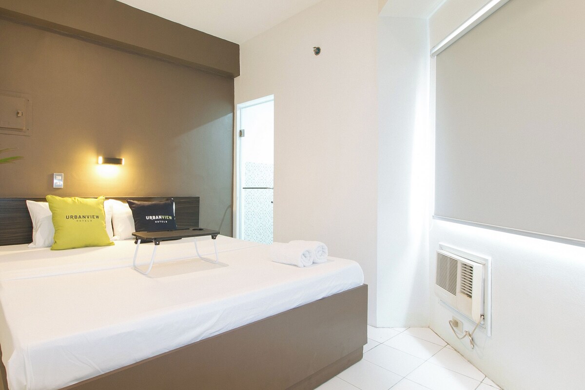 A hotel room with a bed, a bedside table, a potted plant, a wall-mounted TV, and a window.