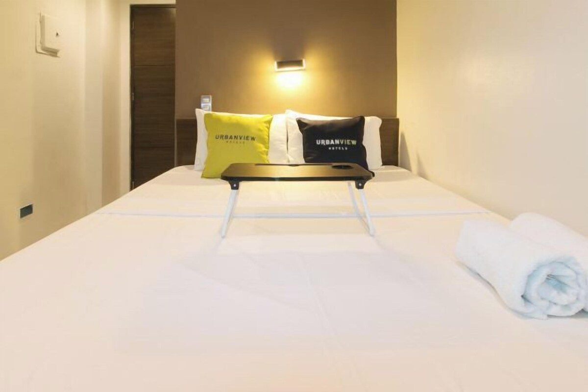 A hotel room with a bed, pillows, a tray table, and a rolled towel.