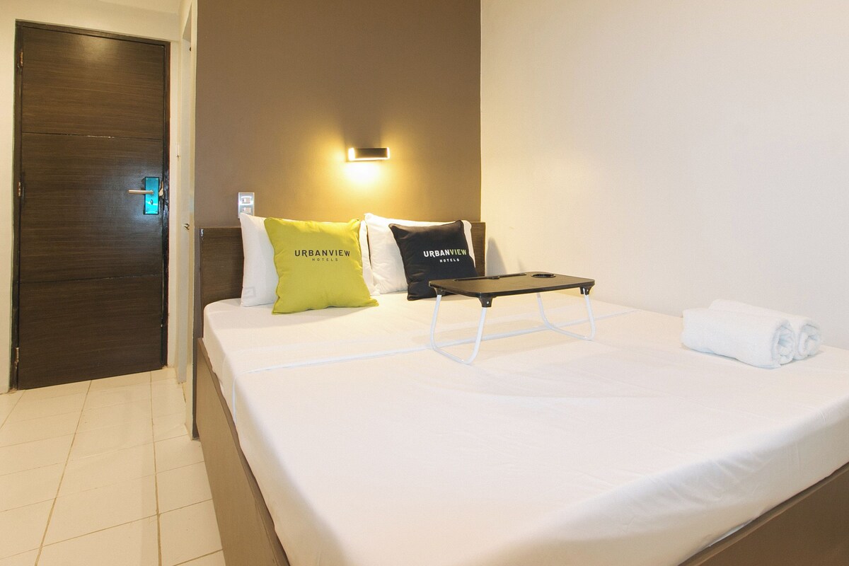 A hotel room with a bed, pillows, a bedside table, and a wall-mounted light.