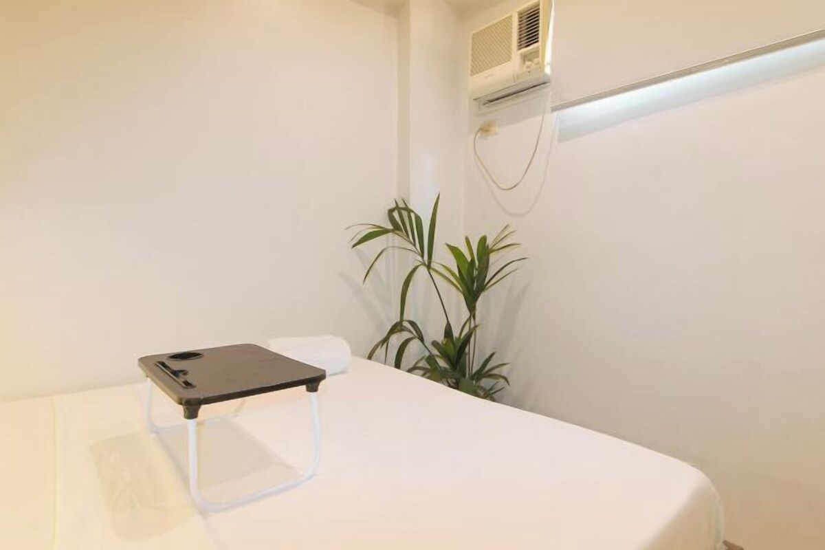 Een nette, minimalistisch ingerichte kamer met een bed, een klein tafeltje met een smartphone, een potplant en een airconditioner aan de muur.