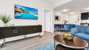 A modern living room with a large flat-screen TV, a dark wooden console table, a blue sofa, and a dining area with blue chairs. - Stylish and New! 3br, Steps fr. Universal Drive (Orlando)