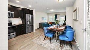A modern kitchen with dark cabinets, stainless steel appliances, and a dining area with blue chairs and a wooden table. - Stylish and New! 3br, Steps fr. Universal Drive (Orlando)