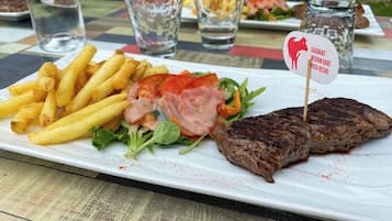 Une assiette contenant un steak, des frites et une salade, avec un verre d’eau et un pot de sauce en arrière-plan.