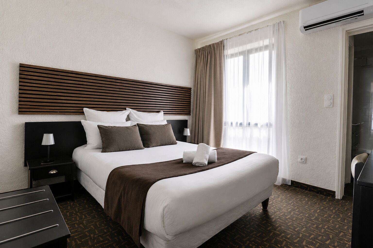 A hotel room with a large bed, a bedside table, a lamp, a window with curtains, and an air conditioning unit.